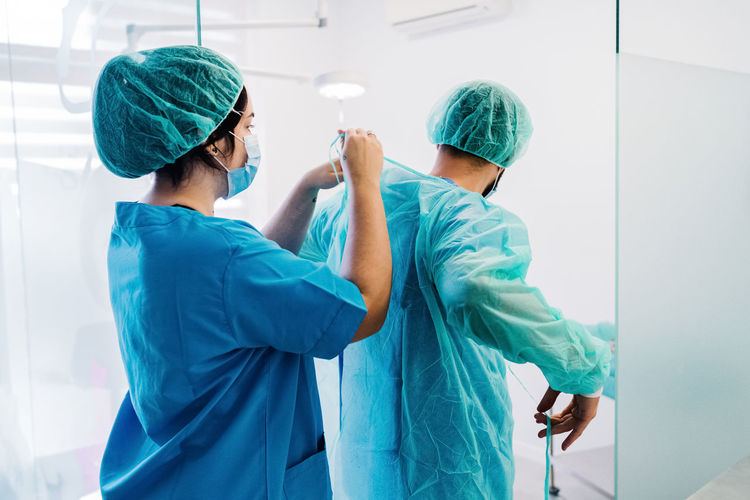Back view of vet doctors putting on protective uniform and gloves while standing in bright operating theater of veterinarian hospital and preparing for surgery