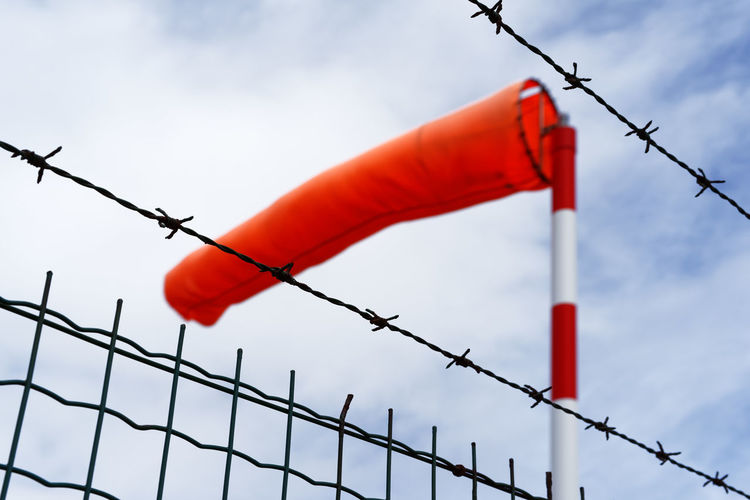 View from below of a bright red bulging windsock of an airfield behind a metal fence