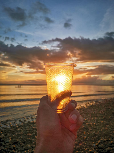 Man holding glass on beach against sky during sunset