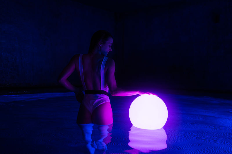 Back view of slim female in swimsuit standing in dark thermal bath pool with luminous ball on water