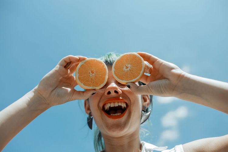 Low angle view of cheerful woman holding orange slices against blue sky