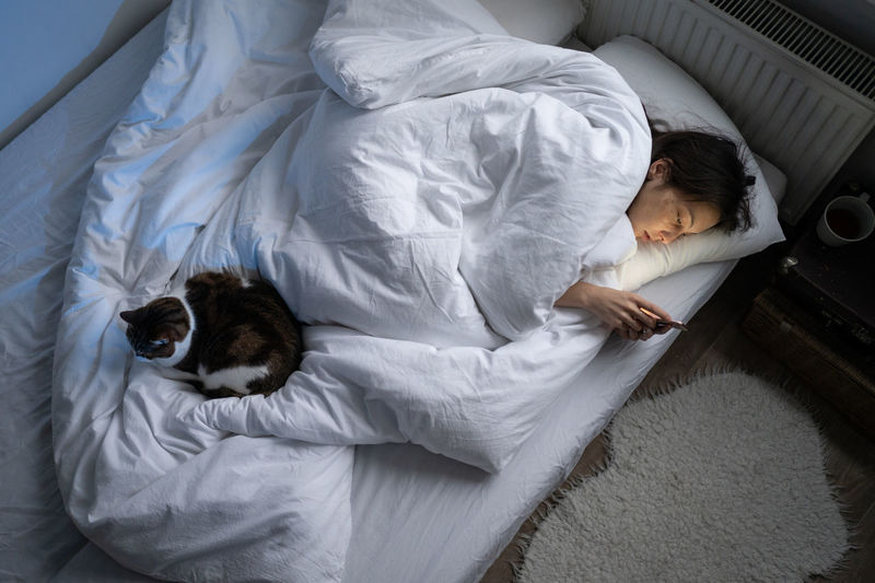 Young lonely depressed woman using smartphone while lying alone in bed with cat in at night