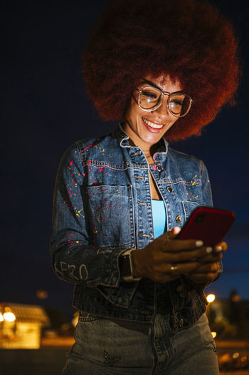 From below positive female with afro hairstyle and modern clothes text messaging on cellphone while walking around the city at night