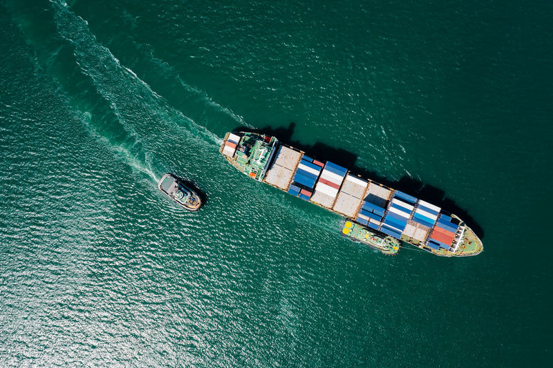 Container ship and tugboat dragging line in green sea aerial top view from drone