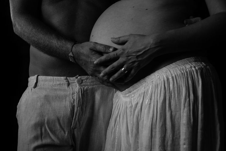 Midsection of shirtless man and woman holding pregnant belly against black background