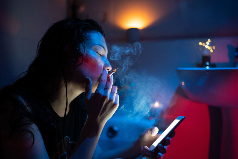 Young woman addicted to nicotine smoking cigarette and using smartphone under neon light in bathroom