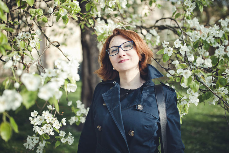 Confedent middle aged armenian woman in a blue trench coat under the blooming tree. spring park 
