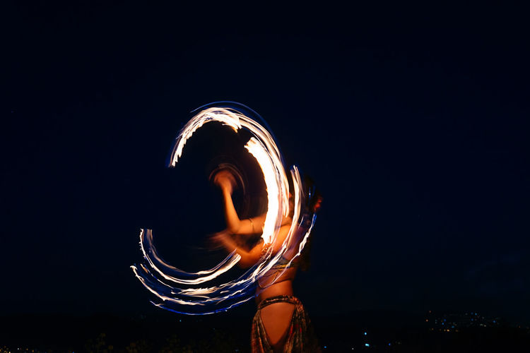 Long exposure of unrecognizable skilled fire dancer with fire fan standing on dark street during performance on night time in city
