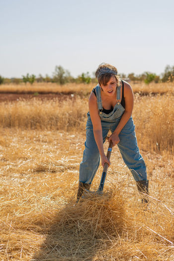 Women in overall and rubber boot using rake for harvesting dried grass in village in summer