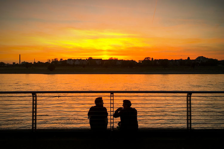 Silhouette men standing on riverbank against sky during sunset