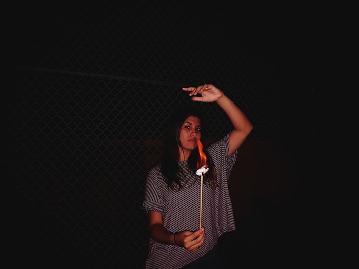 Portrait of woman with burning stick while standing against fence at night 