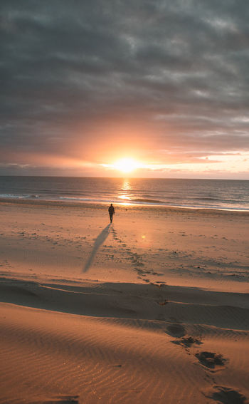 Silhouette man walking at beach against cloudy sky during sunset