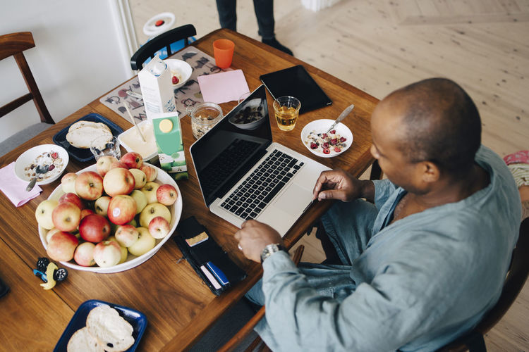 High angle view of mature man shopping online on laptop while having breakfast at table in dining room