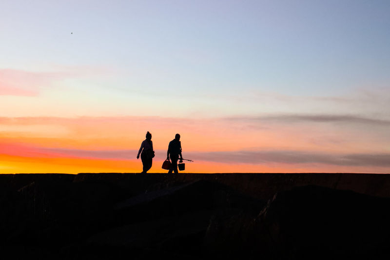 Silhouette people on sidewalk against sky during sunset