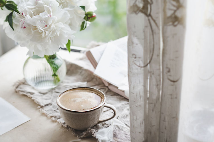 Stylish ceramic cup with  coffee and foam at window with curtains and glass vase peonies, cozy home