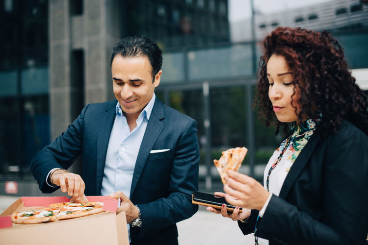 Businessman and businesswoman eating pizza against building during lunch