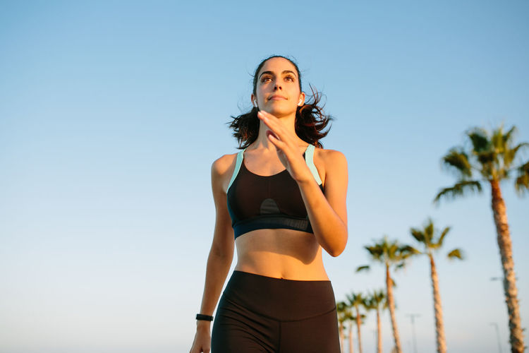 From below of determined young slim female in black sports top and leggings running on street with palm trees against blue sky
