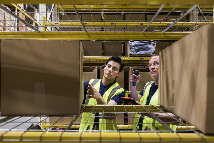 Male coworkers discussing over cardboard box while seen through rack at warehouse