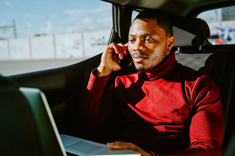 Busy african american businessman sitting in luxury car with laptop and speaking with client on smartphone while looking away