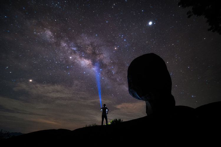 Silhouette man lighting flashlight towards nebula in sky at night