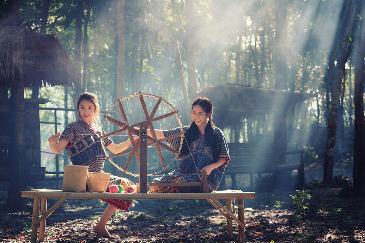 Women using spinning wheel while sitting on wooden bench outdoors
