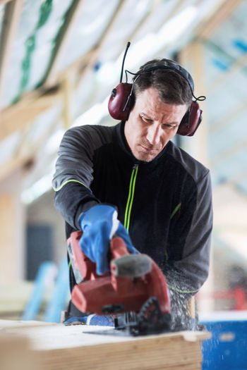 Concentrated manual worker wearing ear protectors sawing wooden planks at site