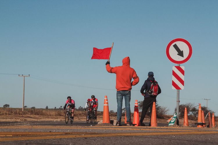 Rear view of man holding red flag while cyclists riding bicycles on road against clear sky