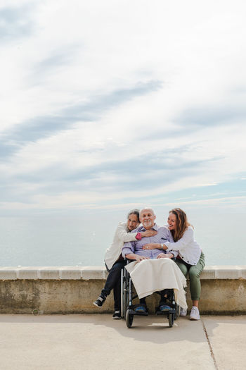 Happy family hugging aged father sitting in wheelchair on embankment near sea on sunny day