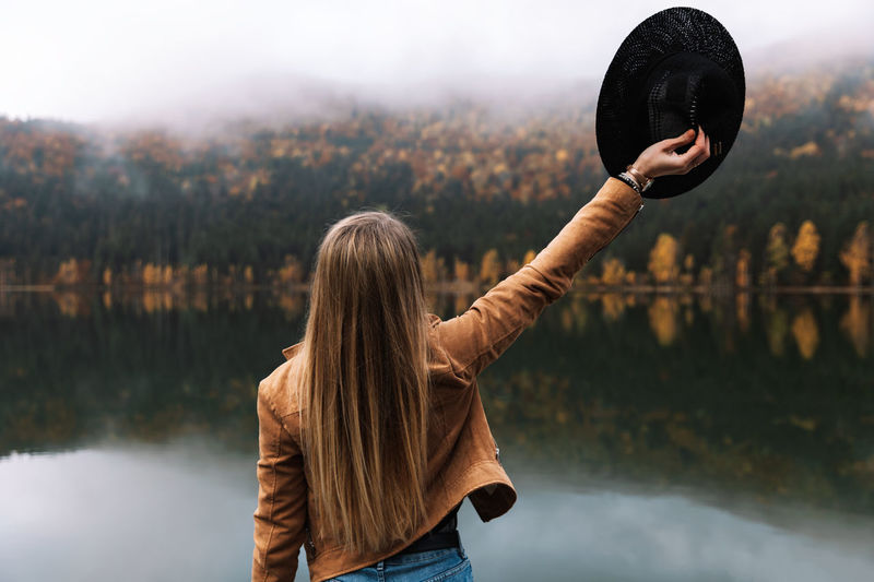 Young woman with raised arm on background of autumn forest and mountain lake. outdoor, rear view.	