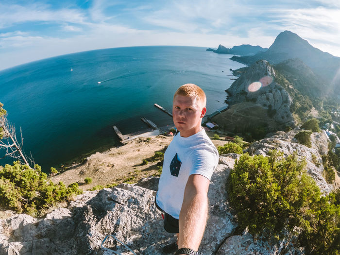 Portrait of young man taking selfie while standing on cliff by sea
