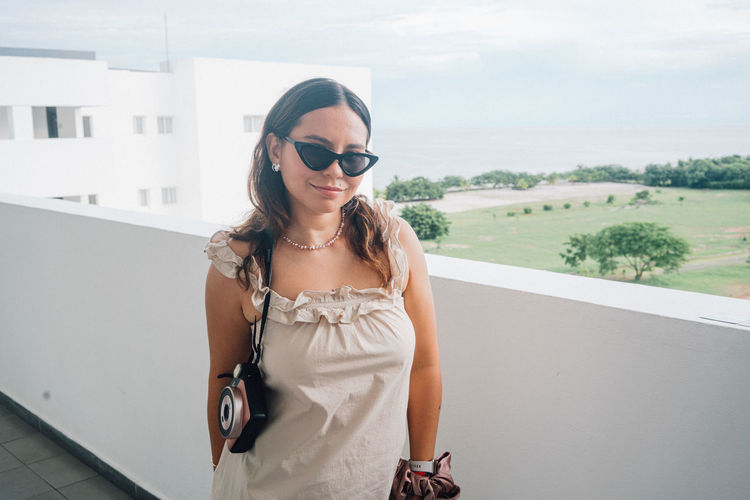Portrait of young woman wearing sunglasses while standing against sea with an instasquare camera 