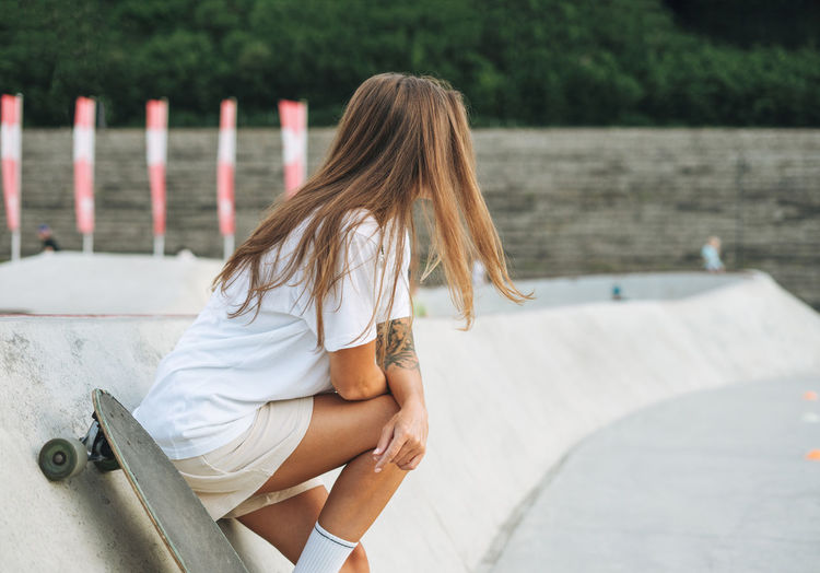Slim young woman with long blonde hair in light sports clothes with longboard in outdoor skatepark 