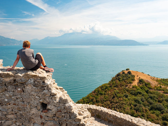 Tired blond hair boy sit on wall of ruin rocca di manerba enjoy view to south garda lake, italy