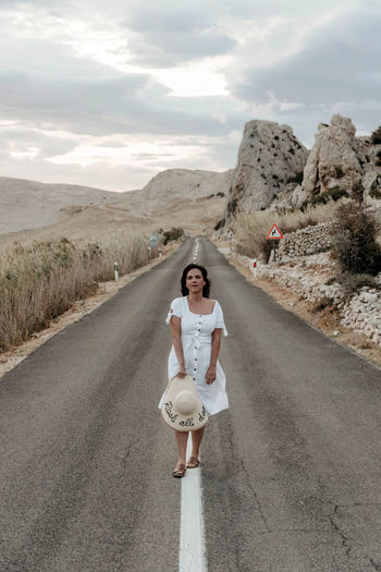 Front view of beautiful young woman in white dress standing on open road.