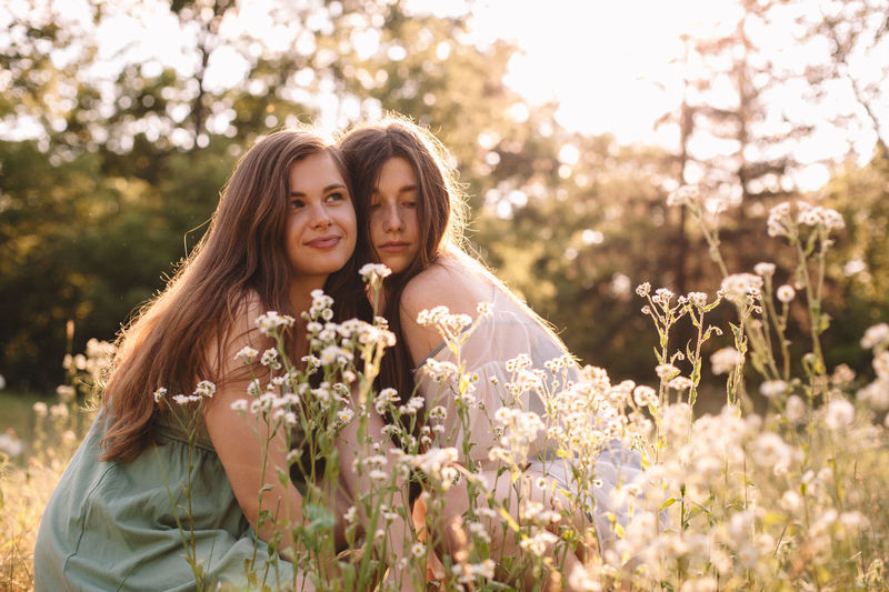Portrait of two girlfriends sitting amidst flowers in summer forest