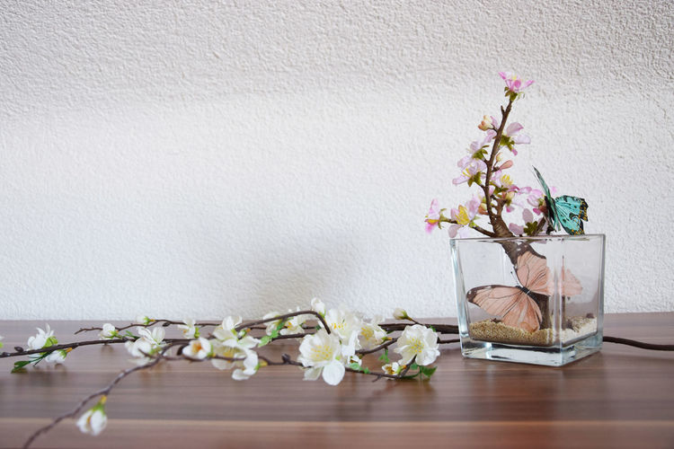 Close-up of ikebana with artificial butterflies on table against white wall