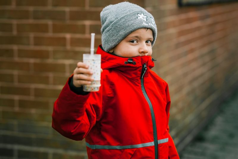 Portrait of boy holding container while wearing red warm clothing against brick wall