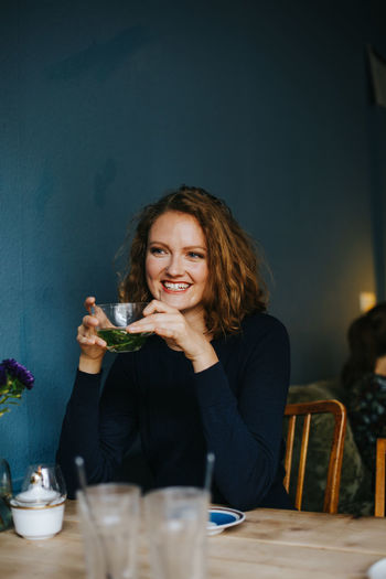 young woman holding cup of tea and smiling Adult Beautiful Woman Cheerful Food Food And Drink Happiness Holding Cup Indoors Night One Person People Portrait Real People Sitting Smiling Smiling Face Table Young Adult