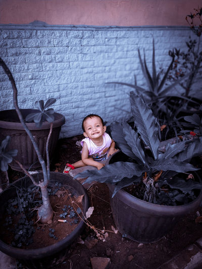 Portrait of smiling boy sitting behind potted plants against wall 