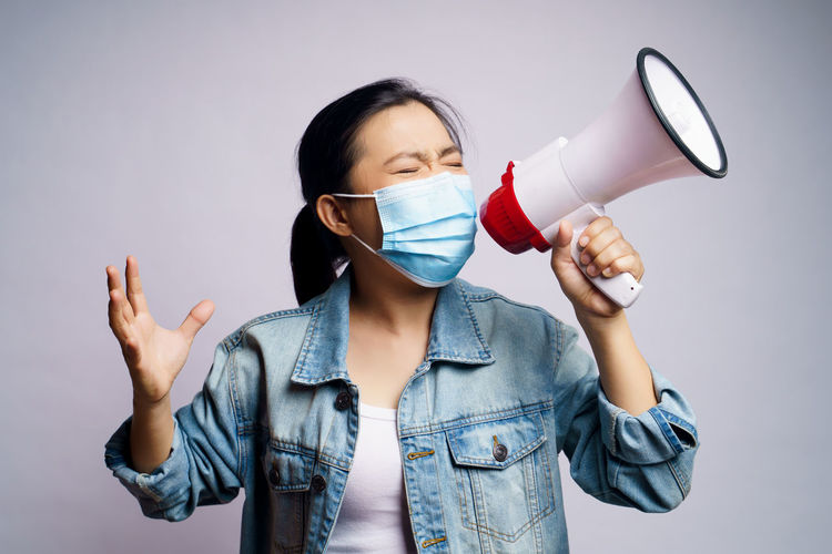 Close-up of young woman wearing flu mask screaming on megaphone against white background