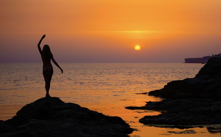 Silhouette woman standing on rock on shore against sky during sunset