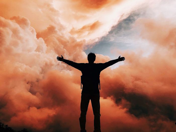 Man with arms outstretched standing against cloudy sky during sunset