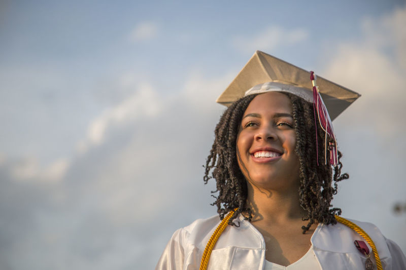 Smiling young woman wearing mortarboard against sky