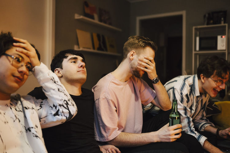 Disappointed man covering eyes while sitting with friends in living room