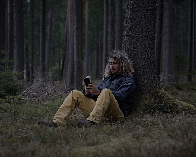 Man holding diary while leaning on tree trunk in forest