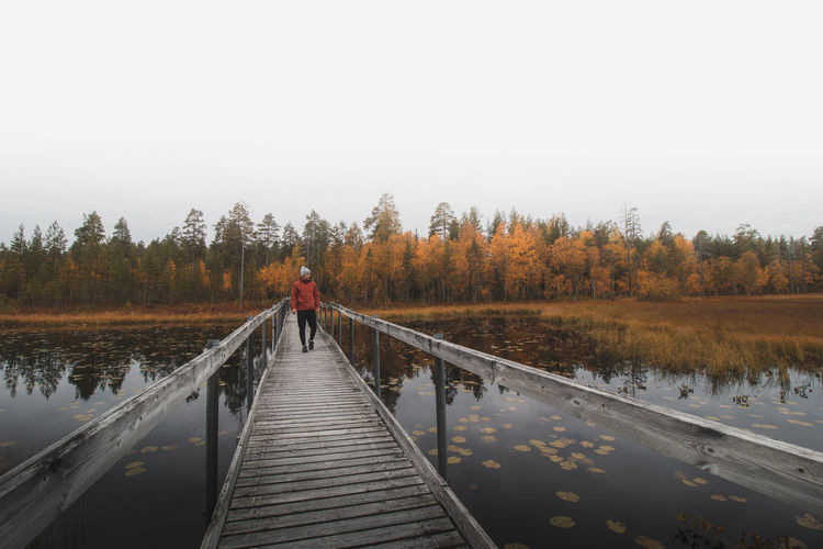 Man in a red jacket and cap walks on a wooden bridge, looking out over lake. kainuu region, finland.