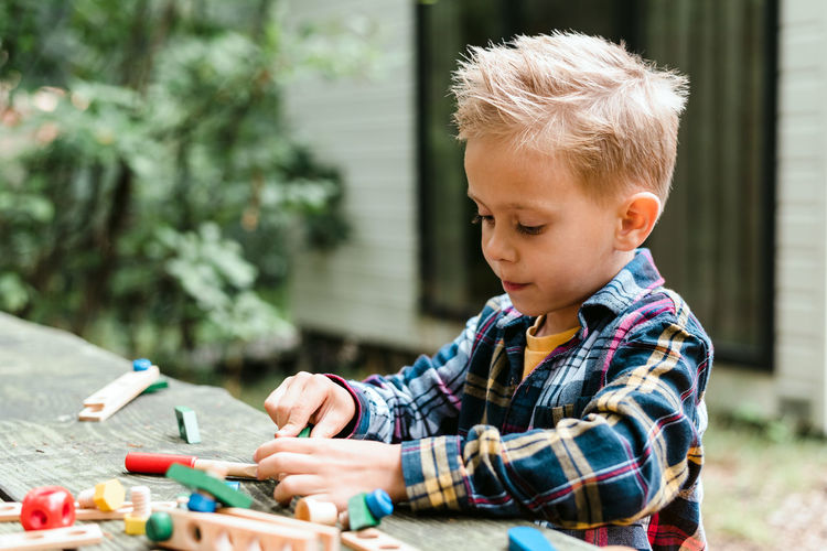 Little boy in a checkered shirt plays with a wooden constructor outdoors.