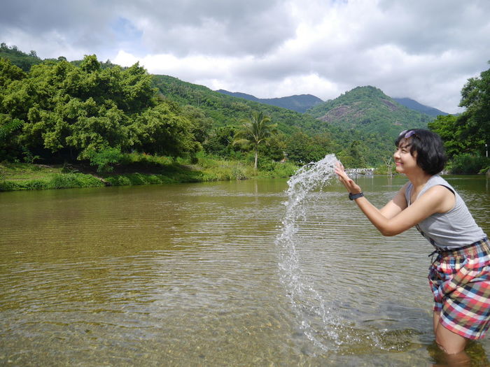Cheerful young woman splashing water in lake against cloudy sky