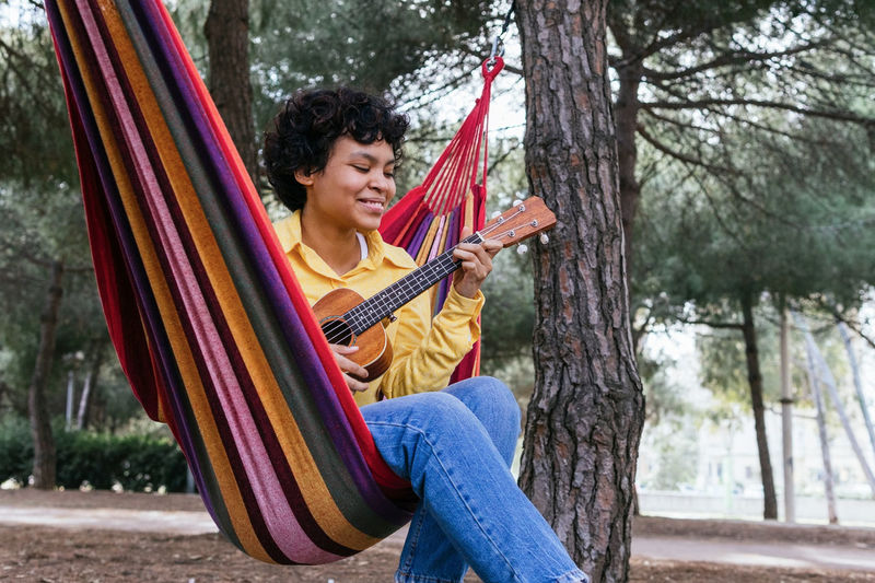 Smiling young ethnic female in bright shirt lounging in park hammock and playing ukulele