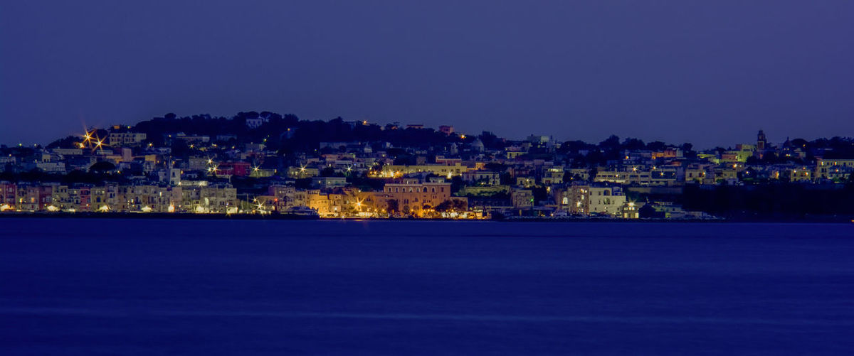 Illuminated buildings by sea against blue sky at night